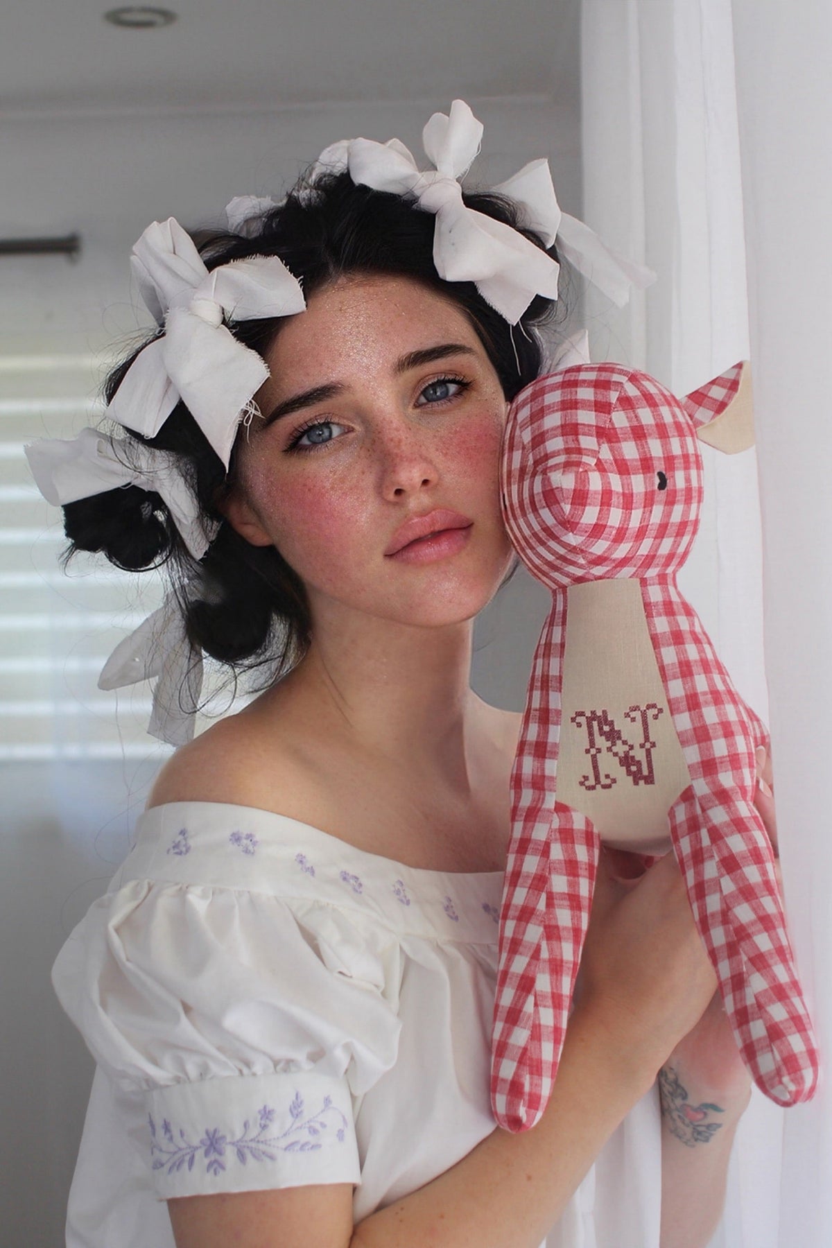 A woman with dark hair styled in a vintage updo with white bows, wearing a white off-the-shoulder blouse with blue floral embroidery, holding a red and white gingham stuffed animal toy with a monogrammed 'N' against a white curtain background.